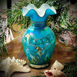 May include: A turquoise glass vase with a ruffled white rim and gold glitter accents. The vase features a seahorse design and a web-like pattern. Decorative seashells and greenery surround the vase.
