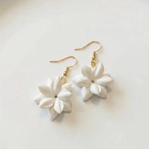 May include: A pair of white flower earrings with gold-colored hooks. The earrings feature detailed, multi-petal flower designs. The earrings are displayed on a white surface.