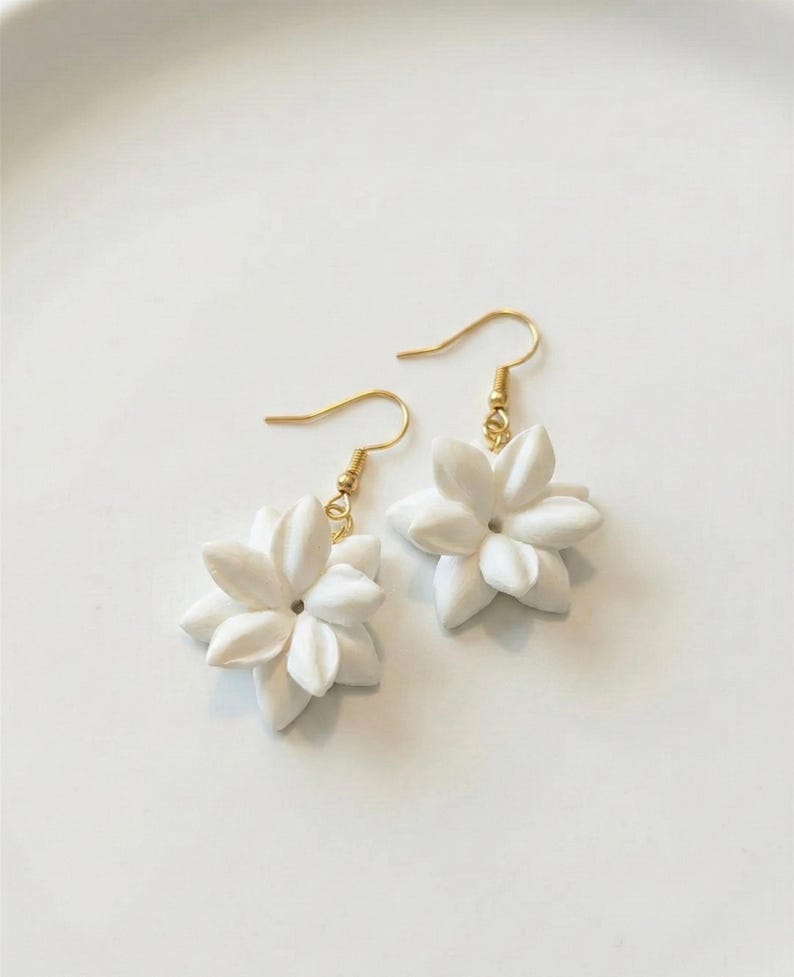 May include: A pair of white flower earrings with gold-colored hooks. The earrings feature detailed, multi-petal flower designs. The earrings are displayed on a white surface.
