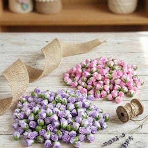 Puede incluir: Dos montones de capullos de rosa artificiales en tonos rosa y lavanda, con acentos verdes, sobre una superficie de madera clara. Una cinta de arpillera y una bobina de hilo de madera sugieren suministros de artesanía para arreglos florales.