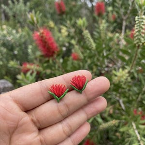 Puede incluir: Un par de pendientes de botón con forma de flores de limpiatubos rojas con hojas verdes. Cada pendiente presenta un grupo de estambres finos de color rojo vibrante que irradian hacia afuera, sostenidos por pequeñas hojas verdes esculpidas.