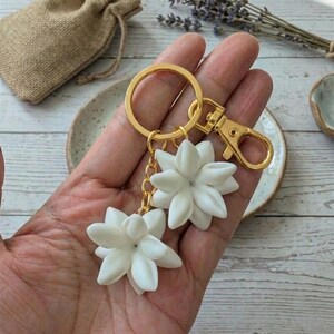 May include: A gold-colored keychain with two white flower charms. The charms are detailed with multiple petals and a small gem in the center. The keychain includes a clasp and a key ring. A small burlap bag is in the background.