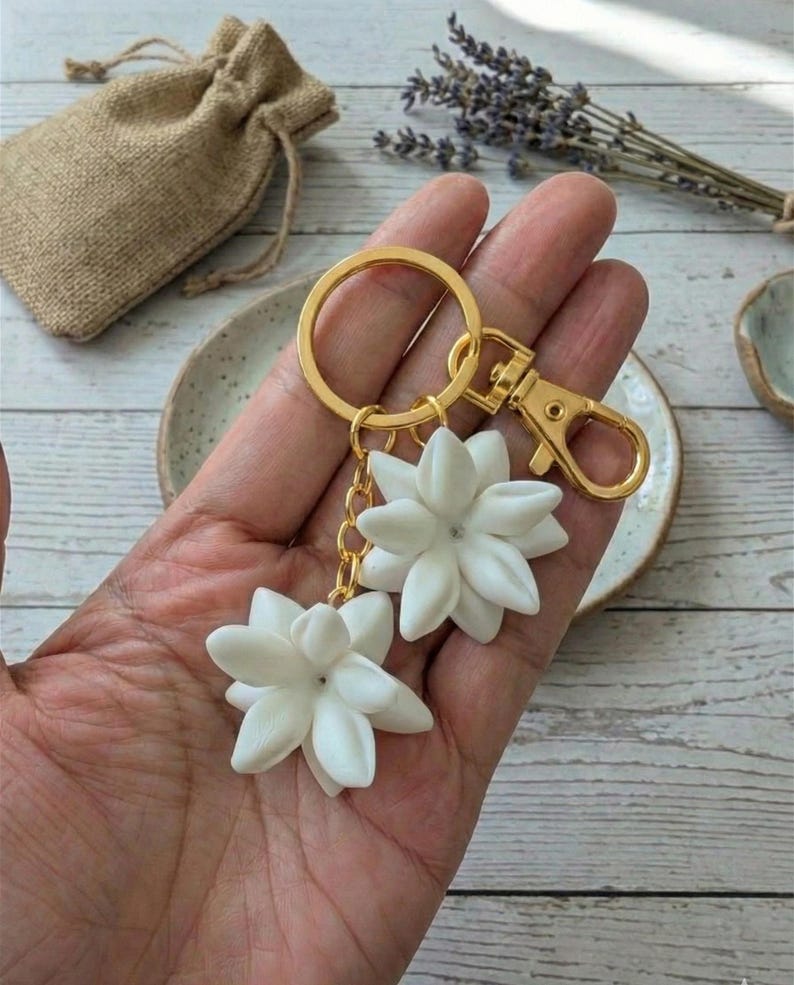 May include: A gold-colored keychain with two white flower charms. The charms are detailed with multiple petals and a small gem in the center. The keychain includes a clasp and a key ring. A small burlap bag is in the background.