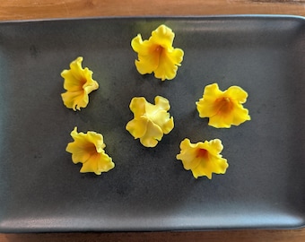 Puede incluir: Un plato rectangular gris oscuro con seis flores amarillas brillantes dispuestas encima. Las flores tienen un diseño de pétalos rizados y un centro amarillo más oscuro. El plato está sobre una superficie de madera.