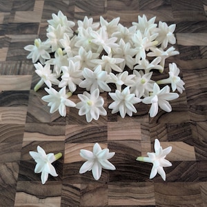 May include: A collection of white jasmine flowers with multiple petals and green stems, arranged on a dark brown wooden surface. The flowers are in various stages of bloom, with some fully open and others still budding.