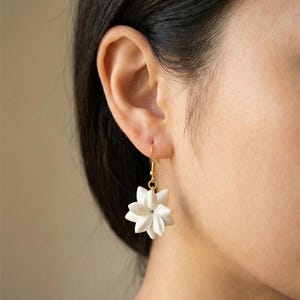 May include: White flower-shaped earrings with gold-colored hooks. The earrings are a delicate, floral design, with multiple petals creating a three-dimensional effect. The earrings are shown against a blurred background.