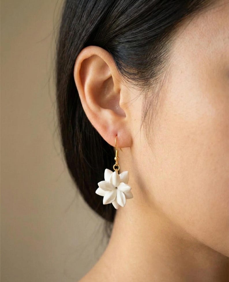 May include: White flower-shaped earrings with gold-colored hooks. The earrings are a delicate, floral design, with multiple petals creating a three-dimensional effect. The earrings are shown against a blurred background.