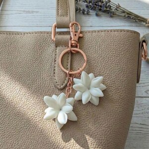 May include: A beige leather tote bag with a rose gold keychain featuring two white flower charms. The bag has a textured surface and a matching handle. Dried lavender sprigs are visible in the background.