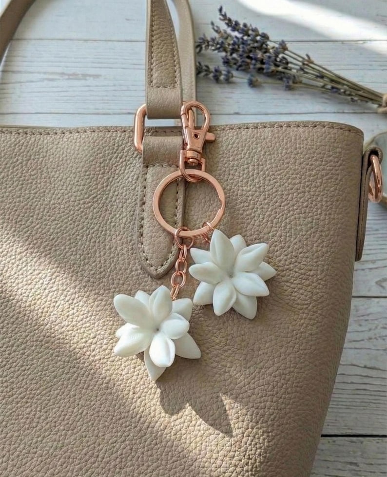 May include: A beige leather tote bag with a rose gold keychain featuring two white flower charms. The bag has a textured surface and a matching handle. Dried lavender sprigs are visible in the background.