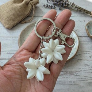 May include: A silver keychain with two white flower charms. The charms are detailed with multiple petals, and are connected to the keychain with a chain. The keychain is held in a hand, with a small burlap bag in the background.
