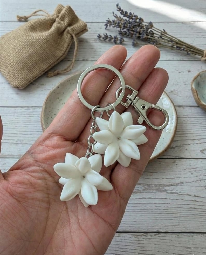 May include: A silver keychain with two white flower charms. The charms are detailed with multiple petals, and are connected to the keychain with a chain. The keychain is held in a hand, with a small burlap bag in the background.