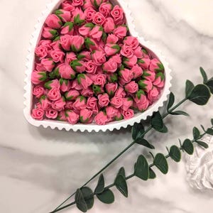 Puede incluir: Un plato blanco en forma de corazón lleno de capullos de rosas rosas. El plato tiene un borde festoneado y está lleno de pequeñas flores. Se ven tallos y hojas verdes cerca del plato, lo que añade un toque natural.