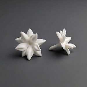 May include: Two white ceramic flower sculptures. One is a fully bloomed flower with multiple petals, and the other is a partially bloomed flower. The sculptures are set against a gray background.