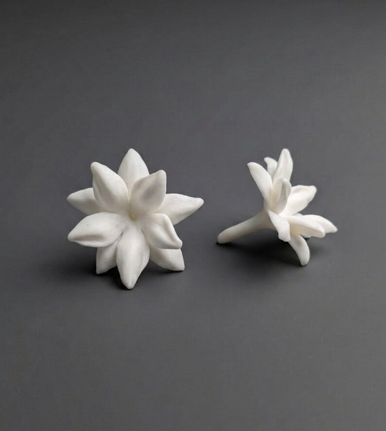 May include: Two white ceramic flower sculptures. One is a fully bloomed flower with multiple petals, and the other is a partially bloomed flower. The sculptures are set against a gray background.