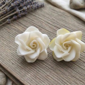 Puede incluir: Dos objetos en forma de rosa de color blanco roto con pétalos curvos y superpuestos. Las flores están colocadas una al lado de la otra y presentan un acabado liso y mate. Cada rosa tiene un centro enrollado que se abre hacia pétalos exteriores.
