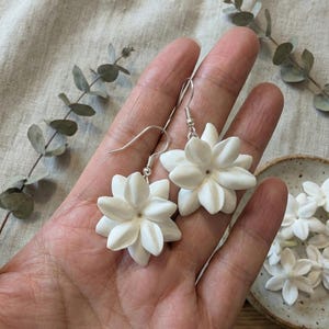 May include: A pair of white flower earrings with silver hooks. The earrings are in the shape of jasmine flowers, with multiple petals. Additional jasmine flower accents are in a small ceramic dish.