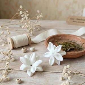 May include: Two white flower-shaped decorations with a central bead are displayed on a light wooden surface. A small terracotta dish with moss, a spool of cream-colored thread, and small beads are also present. The background includes dried flowers and a tag that reads "Etsy - Handmade".