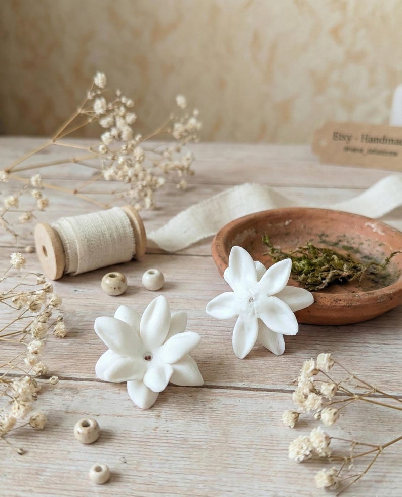 May include: Two white flower-shaped decorations with a central bead are displayed on a light wooden surface. A small terracotta dish with moss, a spool of cream-colored thread, and small beads are also present. The background includes dried flowers and a tag that reads "Etsy - Handmade".