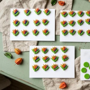 Puede incluir: Varias tarjetas rectangulares blancas con filas de pequeños adornos tridimensionales de flores ave del paraíso en vibrantes tonos naranja, azul y verde. Una tarjeta muestra cuatro formas circulares verdes lisas en lugar de flores.