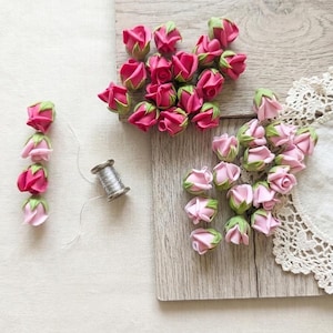 Puede incluir: Una colección de pequeños capullos de rosa artificiales en tonos rosa oscuro y rosa claro, con sépalos verdes. Un carrete de metal plateado con hilo fino se encuentra cerca. Los capullos están dispuestos en grupos y en una línea vertical.