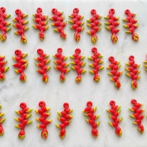Puede incluir: Una colección de veinticuatro pequeños colgantes alargados con forma de flores de heliconia. Cada colgante presenta un esquema de color rojo y amarillo vibrante con un patrón segmentado repetitivo y un pequeño lazo en la parte superior.