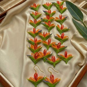Peut inclure: Une collection de petites breloques en forme de fleur oiseau de paradis fabriquées à la main, aux teintes vives orange, jaune et vert, disposées en rangées. En bas, une paire de boucles d'oreilles assorties avec des crochets en métal doré.