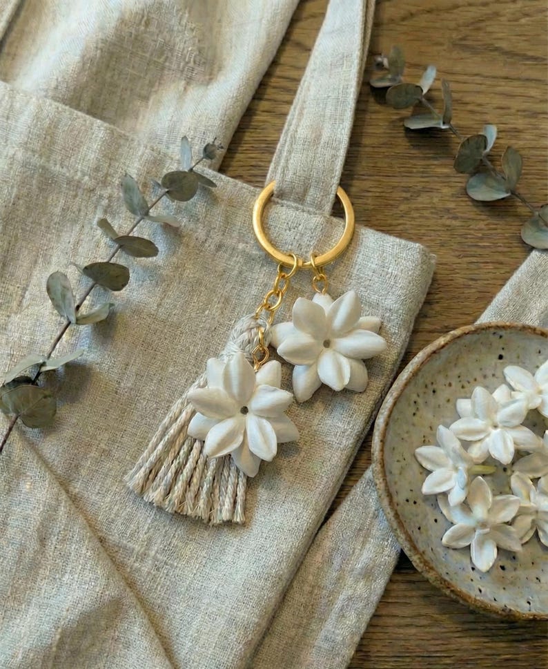 May include: A light gray tote bag with a gold ring and a tassel with two white flower charms. A small ceramic bowl with white flower accents is on the right side of the bag.