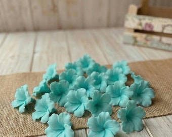 May include: A collection of light blue, delicate flower blossoms. The flowers are clustered on a burlap surface, with a wooden background. The petals are detailed and the overall effect is soft and inviting. The flowers are likely for crafting or decoration.