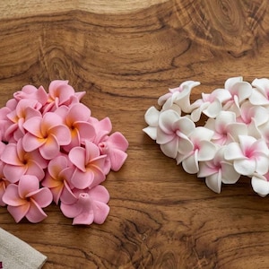 Puede incluir: Dos grupos de flores artificiales de plumeria en rosa y blanco con centros amarillos, dispuestas sobre una superficie de madera. También hay un dije de flor de bronce. Las flores son probablemente para manualidades o decoración.