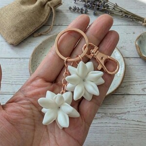 May include: A rose gold keychain with two white flower charms. The charms are detailed with multiple petals and a small gem in the center. The keychain has a clasp and a key ring. A small burlap bag is in the background.