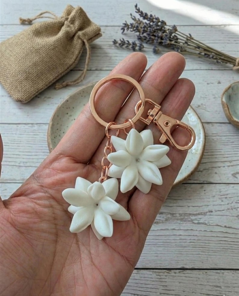 May include: A rose gold keychain with two white flower charms. The charms are detailed with multiple petals and a small gem in the center. The keychain has a clasp and a key ring. A small burlap bag is in the background.