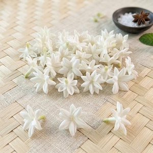 Puede incluir: Un conjunto de pequeñas flores de jazmín blancas con pétalos en forma de estrella dispuestas en grupo. Cerca, un cuenco oscuro contiene cristales de sal blancos, una estrella de anís marrón y una pequeña rama de menta verde.