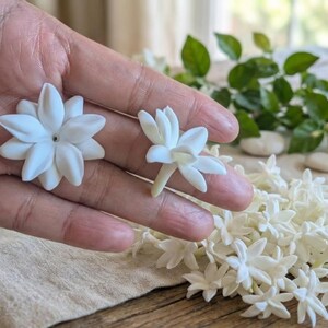 May include: Close-up of white jasmine flowers, showcasing their delicate petals and intricate details. The flowers are arranged on a wooden surface, with a scattering of additional blooms and green foliage in the background. The image highlights the natural beauty of the flowers.