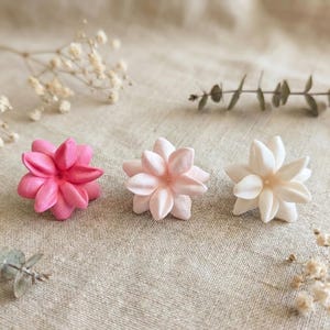 Puede incluir: Tres adornos con forma de flor, en tonos rosa y blanco, dispuestos sobre una tela de tono neutro. Las flores tienen múltiples pétalos y están rodeadas de ramitas verdes y pequeñas flores blancas.