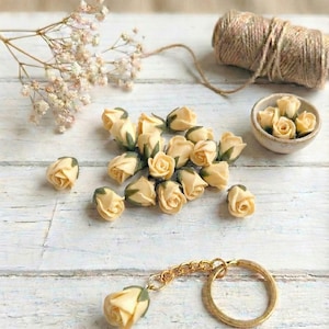 May include: A collection of small, cream-colored rose buds with green accents, scattered on a white wooden surface. A gold-colored keychain with a rose bud charm is in the foreground. A small bowl of rose buds and a spool of twine are also present.