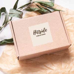 May include: A light brown cardboard box with the words "Hillside Creations" printed on a cream-colored square label. The box is on a bed of brown paper with green leaves in the background.