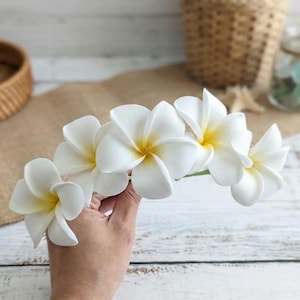 Puede incluir: Un grupo de flores de plumeria blancas con centros amarillos. Las flores se sostienen en una mano, con un fondo borroso que presenta una cesta tejida, un frasco de vidrio y una superficie de arpillera. Ideales para decoración tropical.