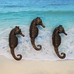 May include: Three bronze-colored seahorse figurines on a white sandy beach with blue ocean waves in the background.
