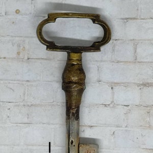 May include: A large, ornate, brass key with a silver-colored shaft and a square-shaped end. The key has a decorative, oval-shaped bow with a cutout in the center.