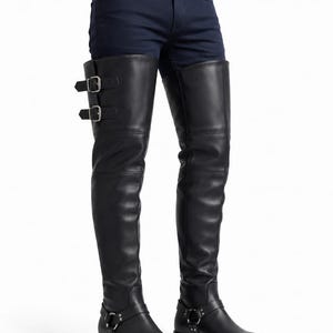 May include: Black leather thigh-high boots featuring a square toe, a low block heel, and a harness strap with a silver-tone metal ring at the ankle. The upper shaft includes two adjustable buckle straps on the outer side of the right boot.