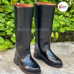 May include: A pair of tall, black leather riding boots. The boots have a smooth, glossy finish and a classic design, with a brown interior. The boots are displayed on a concrete surface, with green foliage in the background. The text "MorarLeatherGear" is visible.