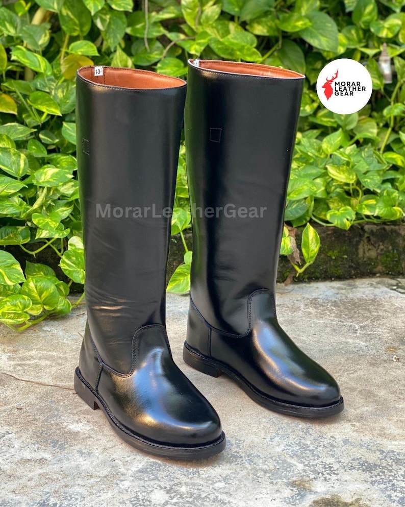 May include: A pair of tall, black leather riding boots. The boots have a smooth, glossy finish and a classic design, with a brown interior. The boots are displayed on a concrete surface, with green foliage in the background. The text "MorarLeatherGear" is visible.