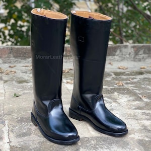 Puede incluir: Un par de botas altas de montar de cuero negro brillante. Las botas tienen un acabado brillante y un interior de color beige. Las botas están sobre una superficie de hormigón. El texto "MorarLeatherGear" es visible.