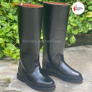 May include: A pair of tall, black leather riding boots. The boots have a smooth, glossy finish and a classic design, with a rounded toe and a flat sole. The brand name "MorarLeatherGear" is visible on the boots.