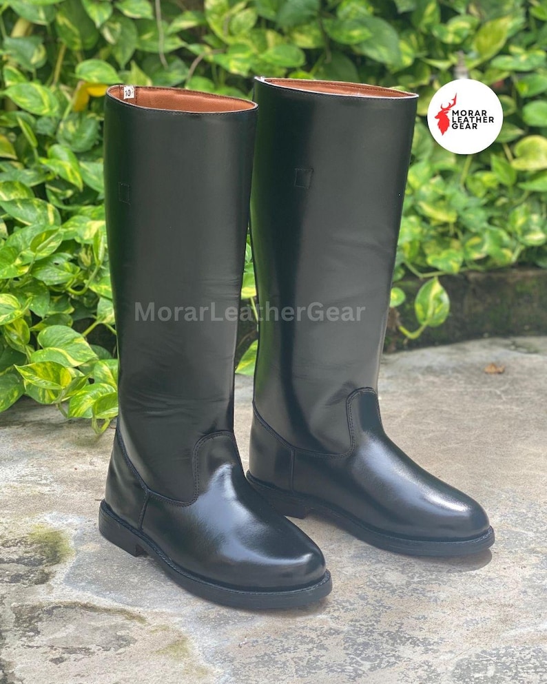 May include: A pair of tall, black leather riding boots. The boots have a smooth, glossy finish and a classic design, with a rounded toe and a flat sole. The brand name "MorarLeatherGear" is visible on the boots.