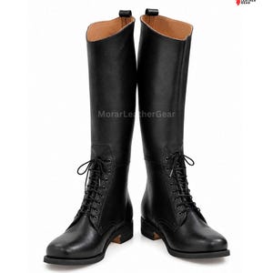 Könnte beinhalten: Ein Paar kniehohe schwarze Stiefel mit Schnürung vorne, Metallösen, abgerundeter Zehenpartie und flachem Absatz. Das Innenfutter ist hellbraun, und der Schriftzug MorarLeatherGear ist auf den Stiefeln sowie oben rechts zu sehen.