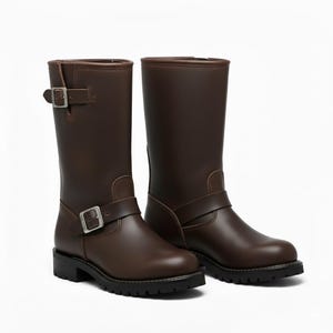 Puede incluir: Un par de botas de moto de cuero marrón. Las botas son de media caña, con punta redonda y suela negra. Cada bota tiene una correa con hebilla alrededor del tobillo y otra en la parte superior. Las botas son de cuero liso y pulido.