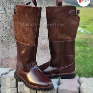 Puede incluir: Un par de botas de cuero marrón. Las botas tienen una parte inferior lisa y brillante y una parte superior de ante. Las botas tienen un cierre de hebilla y una lengüeta en la parte superior. El texto "MorarLeatherGear" es visible.