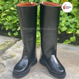 May include: A pair of tall, black leather riding boots. The boots have a smooth, glossy finish and a brown interior. The soles are black and the boots are standing on a concrete surface. The brand name "MorarLeatherGear" is visible.