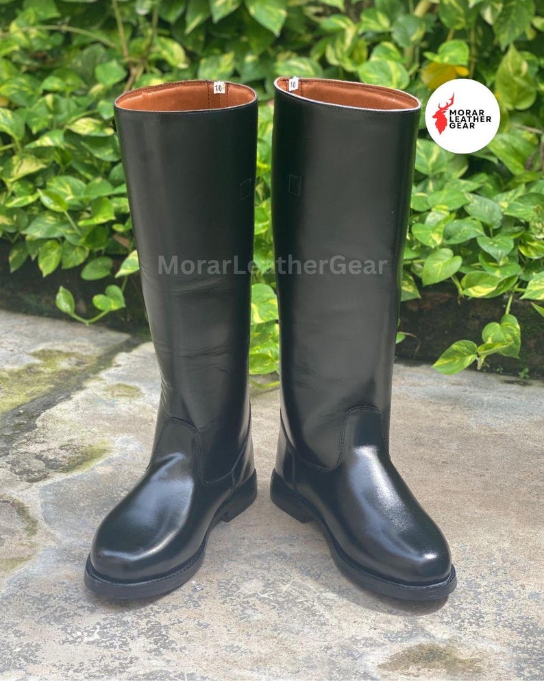 May include: A pair of tall, black leather riding boots. The boots have a smooth, glossy finish and a brown interior. The soles are black and the boots are standing on a concrete surface. The brand name "MorarLeatherGear" is visible.